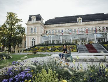 Para siedzi na ławce przed wielkim zabytkowym budynkiem, świętując Europejską Stolicę Kultury 2024.