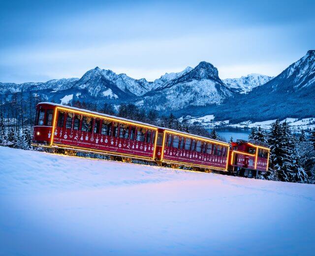 Czerwony pociąg Schafbergbahn ze świecącymi światłami jedzie przez zaśnieżone góry o zmierzchu, wśród sosen.
