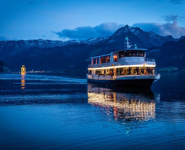 Oświetlony prom płynie o zmierzchu po jeziorze Wolfgangsee, a w tle świecą góry i adwent nad jeziorem.