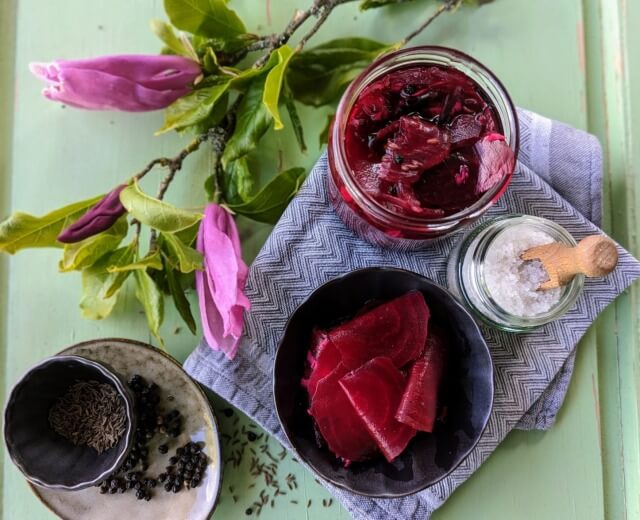 Słoik z buraczkami marynowanymi, słoik z solą, przyprawy i kwiaty ułożone na zielonej tacy z szarą serwetką.