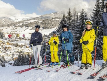 Pięć osób w sprzęcie narciarskim stoi na zaśnieżonym stoku Filzmoos z austriacką wioską i górami w tle.