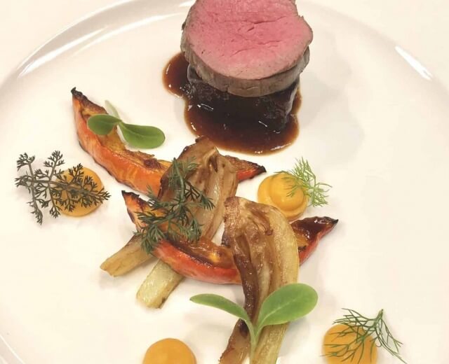 Młoda wołowina polędwica z sosem, pieczonymi warzywami i dodatkami ziołowymi na białym talerzu.