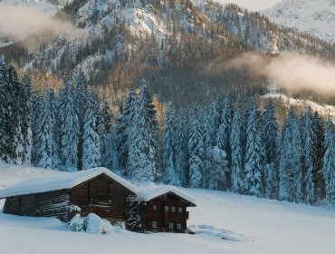 Dwa drewniane domki na zaśnieżonej polanie Zima w Wagrain-Kleinarl otaczają sosny i białe góry.