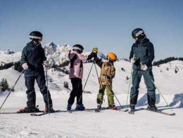 familienskitag-skifahren Cztery osoby w sprzęcie narciarskim cieszą się ferie z rodziną St. Johann na ośnieżonym stoku z widokiem na góry.
