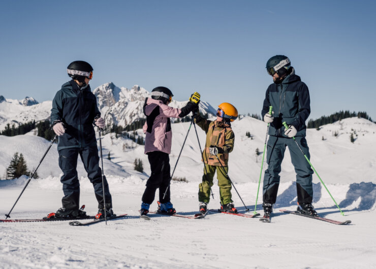 familienskitag-skifahren Cztery osoby w sprzęcie narciarskim cieszą się ferie z rodziną St. Johann na ośnieżonym stoku z widokiem na góry.
