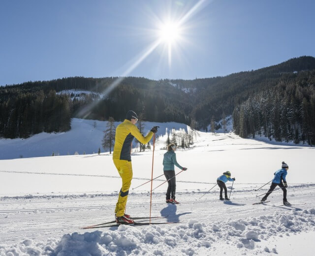 Filzmoos_Langlaufzentrum mit Schneegarantie von Weihnachten bis Ostern©LorenzMasser_www.filzmoos.at Cztery osoby cieszą się narciarstwem biegowym Filzmoos na zaśnieżonym szlaku pod jasnym słońcem i zalesionymi wzgórzami.
