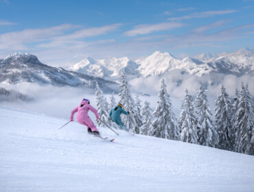 Dwóch narciarzy w kolorowych strojach zjeżdża po zaśnieżonym stoku na nartach w St. Johann, z sosnami i górami za plecami.
