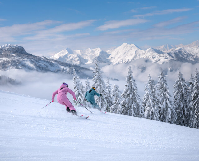 Dwóch narciarzy w kolorowych strojach zjeżdża po zaśnieżonym stoku na nartach w St. Johann, z sosnami i górami za plecami.