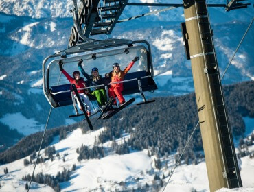 Trzy osoby w sprzęcie narciarskim jadą wyciągiem krzesełkowym nad zaśnieżonymi górami w Narty w Wagrain-Kleinarl, uśmiechając się i machając.