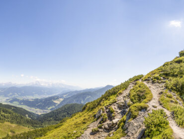 Szczyt górski z krzyżem w pobliżu Flachau, kręty szlak, zielone wzgórza i odległe góry pod czystym, błękitnym niebem.