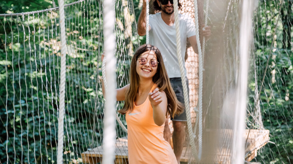 Uśmiechnięta kobieta na moście linowym wskazuje na kamerę, ciesząc się sportem i zabawą St. Johann w leśnej scenerii. (Powiększony widok)