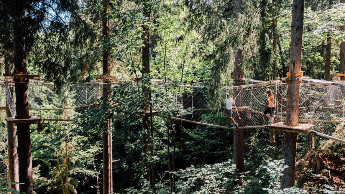 Dwie osoby uprawiają sport i bawią się w St. Johann na moście linowym w słonecznym leśnym parku rozrywki. (Powiększony widok)