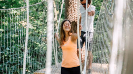 Dwie osoby przechodzą przez most linowy w lesie, uśmiechając się i ciesząc się sportem i zabawą St. Johann.