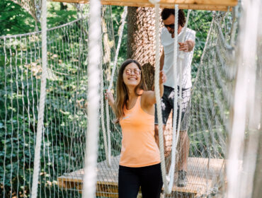 Dwie osoby przechodzą przez most linowy w lesie, uśmiechając się i ciesząc się sportem i zabawą St. Johann.