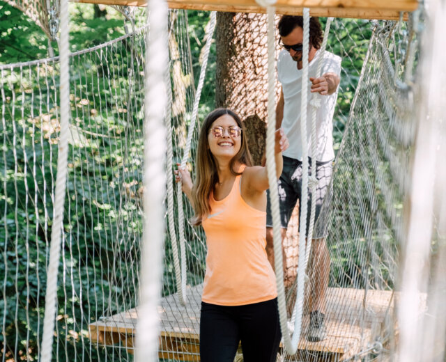 21_st_johann_in_salzburg_sommer_baumparcours_klettern_3 Dwie osoby przechodzą przez most linowy w lesie, uśmiechając się i ciesząc się sportem i zabawą St. Johann.