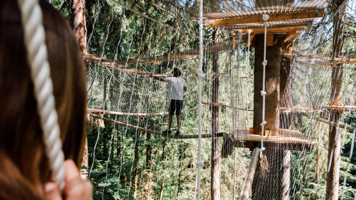 Osoba spacerująca po moście linowym w leśnym parku rozrywki Sport i Zabawa St. Johann, widziana od tyłu. (Powiększony widok)