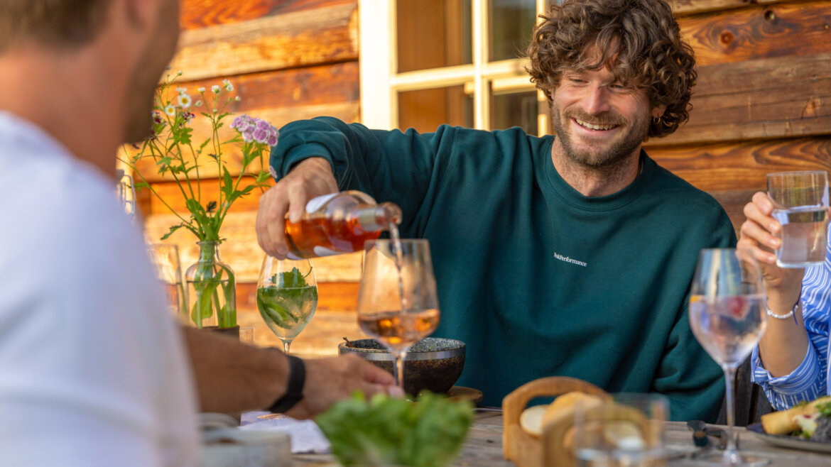 Mężczyzna nalewający drinka przy stoliku na świeżym powietrzu z przyjaciółmi, uśmiechnięty, cieszący się atrakcjami kulinarnymi Flachau.