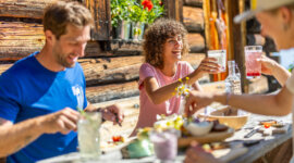 Trzy osoby cieszące się atrakcjami kulinarnymi Flachau przy drewnianym stole na świeżym powietrzu w słoneczny dzień.
