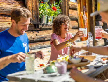 25-flachau-sommer-wandern-berge-25 Trzy osoby cieszące się atrakcjami kulinarnymi Flachau przy drewnianym stole na świeżym powietrzu w słoneczny dzień.