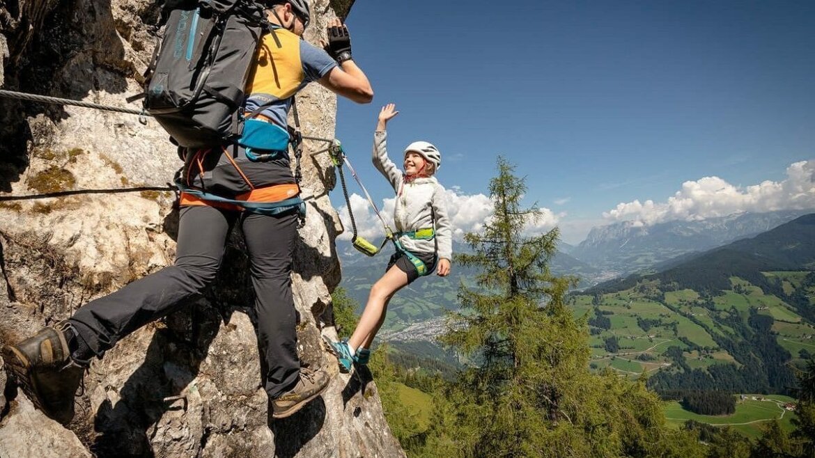 Dwóch wspinaczy przybija piątkę na skalistym klifie z widokiem na kolejki górskie St. Johann i bujne doliny poniżej.
