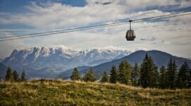 Kolejka linowa przesuwa się po trawiastych wzgórzach, idealnych na turystykę pieszą St. Johann, z ośnieżonymi górami za nią.