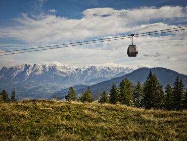 Gondel-Natur Kolejka linowa przesuwa się po trawiastych wzgórzach, idealnych na turystykę pieszą St. Johann, z ośnieżonymi górami za nią.