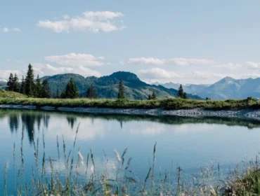 speicherteich-sonntagskogel-alpendorf-1-1920×658.jpg Spokojne jezioro w St. Johann lato, otoczone trawą, drzewami i górami pod błękitnym niebem.