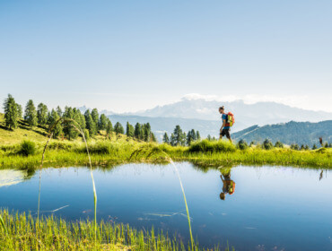 Turysta z plecakiem wędruje brzegiem górskiego jeziora. Wokół zieloe pastwiska, a w tle alepjskie szczyty.