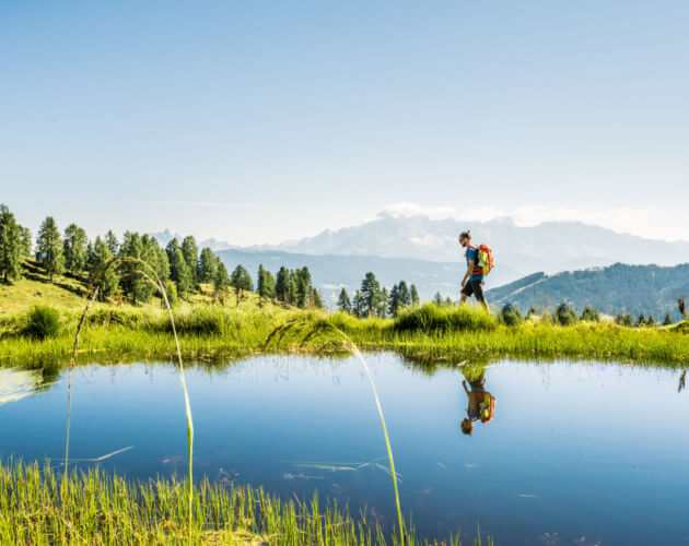 Turysta z plecakiem wędruje brzegiem górskiego jeziora. Wokół zieloe pastwiska, a w tle alepjskie szczyty.