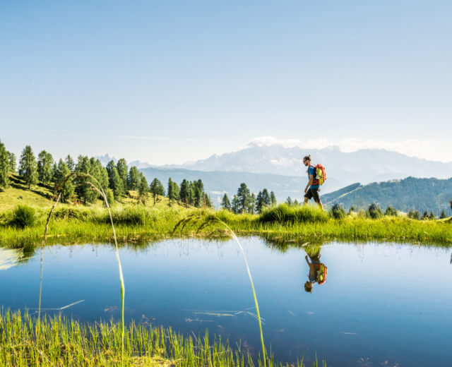 wandern-lackenalmen-in-altenmarkt-zauchensee Turysta z plecakiem wędruje brzegiem górskiego jeziora. Wokół zieloe pastwiska, a w tle alepjskie szczyty.