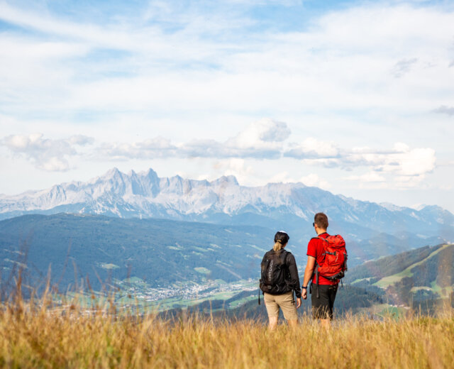 17.08.2022_Grießenkar-sundowner-hike © GO-027 Dwóch wędrowców z plecakami stoi na trawiastym polu, podziwiając Flachau turystyka piesza i odległe górskie widoki.
