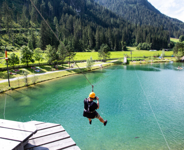 Klettern im Hochseilgarten Flachauwinkl und Flying Fox über den See. Osoba korzystająca z adrenaliny ziplining nad czystym, błękitnym jeziorem we Flachau, w otoczeniu zielonych drzew i gór.