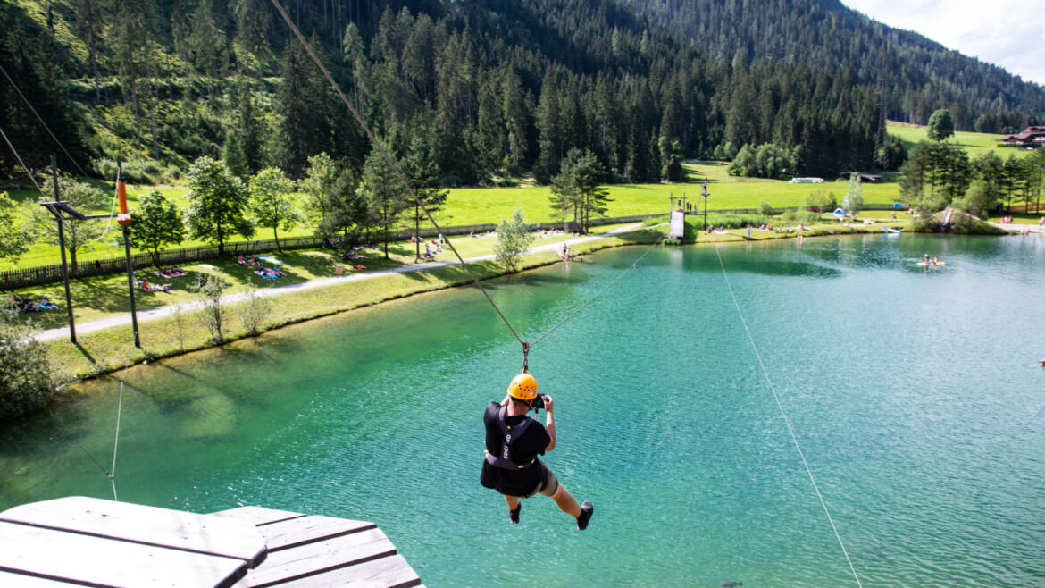 Osoba uprawiająca pełen adrenaliny sport ziplining nad niebiesko-zielonym jeziorem w pobliżu Flachau, w otoczeniu drzew i gór. (Powiększony widok)