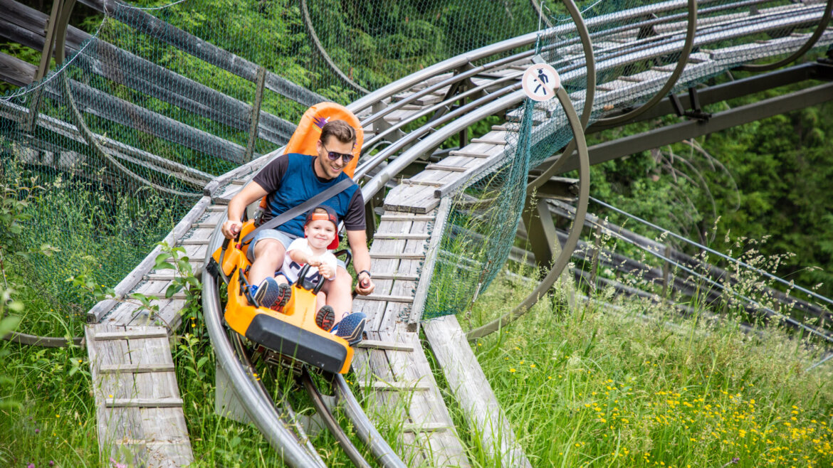Mężczyzna i dziecko cieszą się sportową zabawą na żółtej alpejskiej kolejce górskiej we Flachau, ścigając się wśród trawy i dzikich kwiatów. (Powiększony widok)