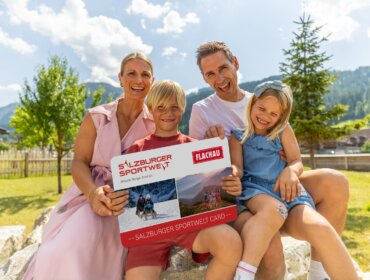 Uśmiechnięta czteroosobowa rodzina trzyma dużą kartę Flachau Sommer Card na zewnątrz w słoneczny dzień.