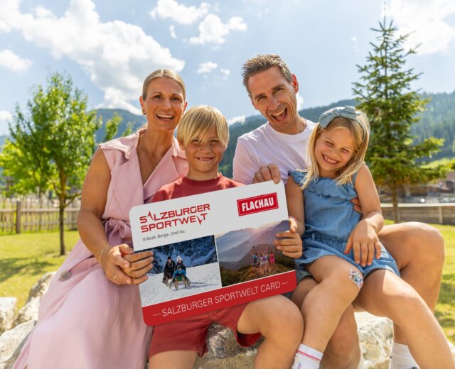 Uśmiechnięta czteroosobowa rodzina trzyma dużą kartę Flachau Sommer Card na zewnątrz w słoneczny dzień.