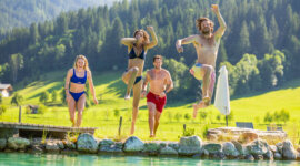 25-flachau-sommer-baden-badeteich-1 Cztery osoby w strojach kąpielowych wskakują do czystego stawu, ciesząc się sportową zabawą i adrenaliną w pobliżu Flachau.