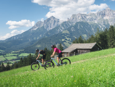 biken-region hochkoeing (C)hochkoenig tourismus Dwóch rowerzystów cieszy się wycieczkami rowerowymi bez bagażu Hochkönig na trawiastym wzgórzu z górami i domkiem w tle.
