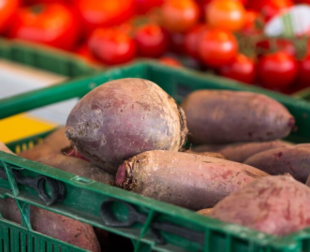 Zielona skrzynia wypełniona burakami na targu, podkreślająca sezon na buraki, z rozmytymi pomidorami z tyłu.