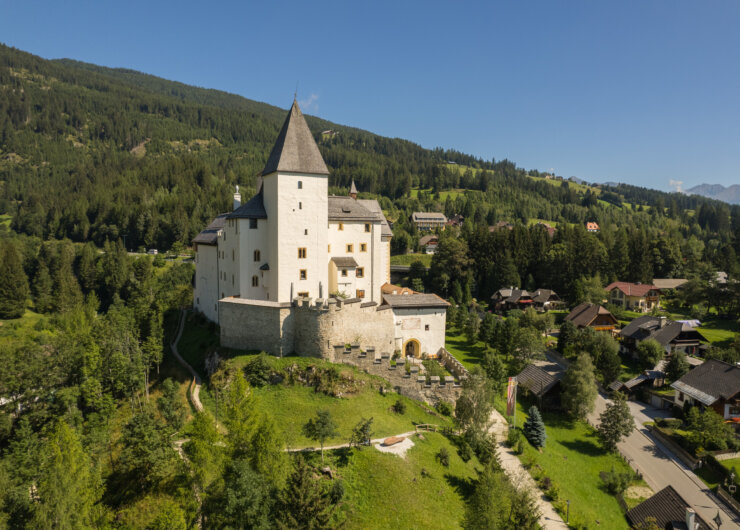 BurgMauterndorf_Aussen_79 – Kopie Biały średniowieczny zamek z wysoką wieżą stoi na zielonym wzgórzu, otoczony drzewami i górami.