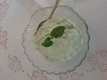 Flaumige-Sauerampfercreme_Lungauer-Kochwerk Szklana miska z kremowym zielonym dipem, prawdopodobnie kremem szczawiowym, jest przyozdobiona bazylią i łyżką w środku.