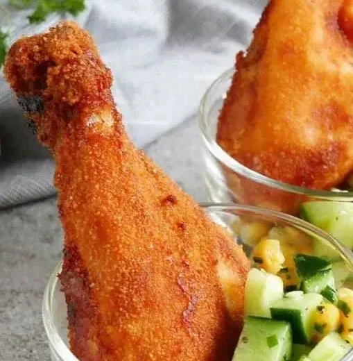 Dwa chrupiące panierowane udka kurczaka podawane w szklanych miseczkach z sałatką ze świeżego ogórka i kukurydzy.