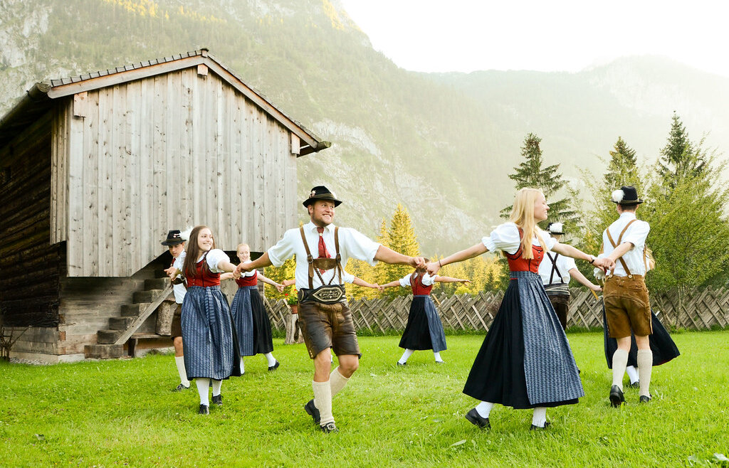 Ludzie w tradycyjnych austriackich strojach tańczący w Tennengau w pobliżu drewnianego budynku i gór. (Powiększony widok)