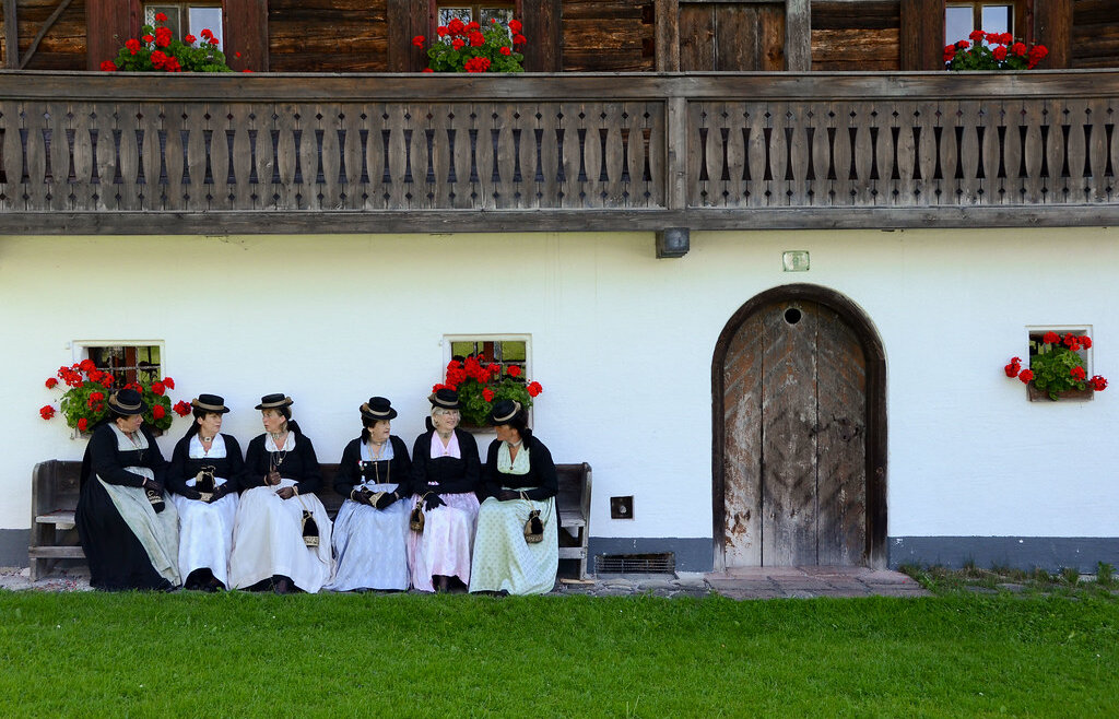 Sześć kobiet w tradycyjnych strojach Tennengau siedzi na ławce przed rustykalnym budynkiem z czerwonymi kwiatami w oknach. (Powiększony widok)