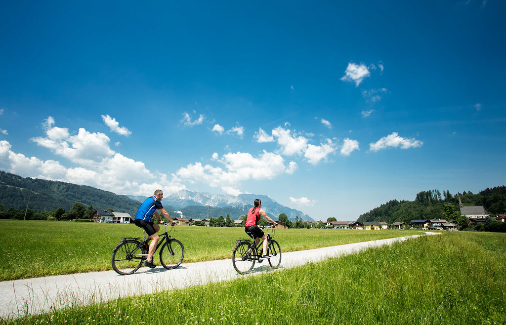 Dwie osoby jadą na rowerach ścieżką przez zielone pola Tennengau z górami i błękitnym niebem za nimi.