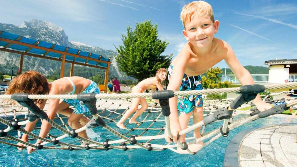 Trójka dzieci wspina się po moście linowym nad basenem w Tennengau z górami i drzewami w tle.