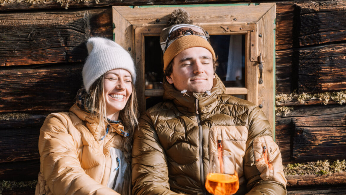 Dwie osoby w zimowych kurtkach relaksują się w drewnianym domku, delektując się Flachau poza stokami z pomarańczowymi napojami. (Powiększony widok)