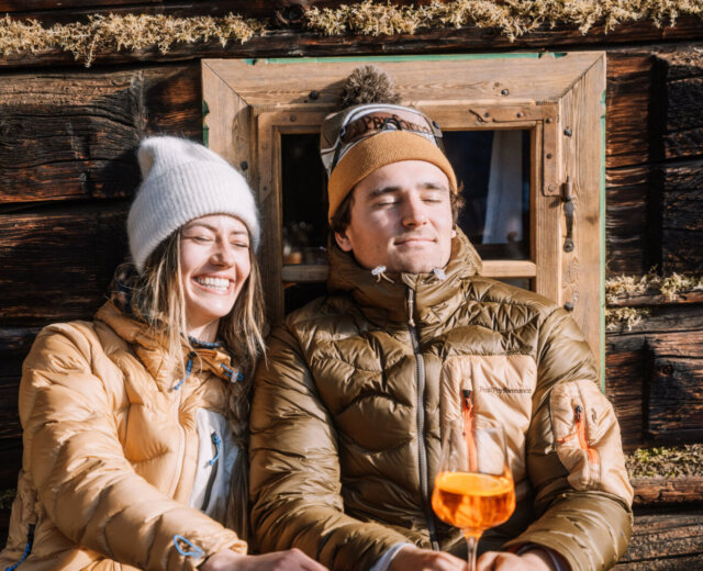 flachau-enjoyment-hut-1600×1067 (1) Dwie osoby w zimowych kurtkach relaksują się w drewnianym domku, delektując się Flachau poza stokami z pomarańczowymi napojami.