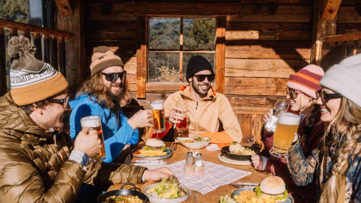 Pięciu przyjaciół w zimowych ubraniach śmieje się i pije drinki we Flachau poza stokami przy słonecznym stoliku na świeżym powietrzu. (Powiększony widok)