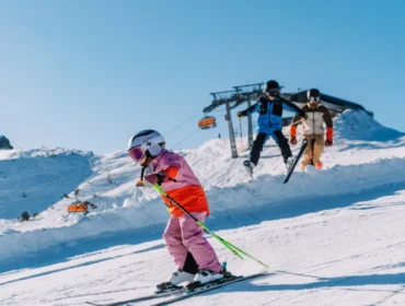 flachau-skiing-family-1170×658.jpg Dziecko w różowych nartach zjeżdża z góry we Flachau ferie z rodziną, dorośli podążają pod czystym, błękitnym niebem.